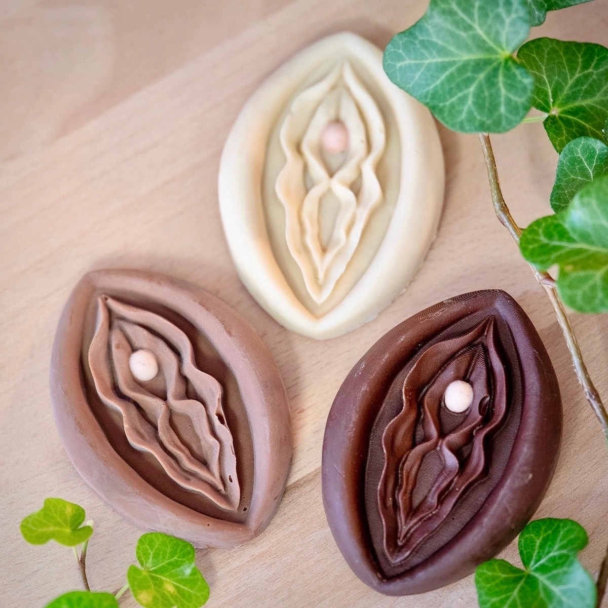 ChocoVulves artisanales en chocolat – Delémont, Suisse Trois ChocoVulves en chocolat noir, au lait et blanc disposées sur du bois avec des feuilles de lierre